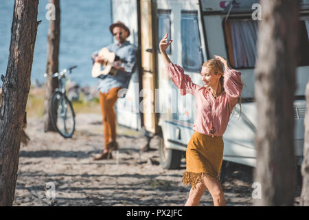 young hippie woman dancing while man playing guitar near trailer Stock Photo