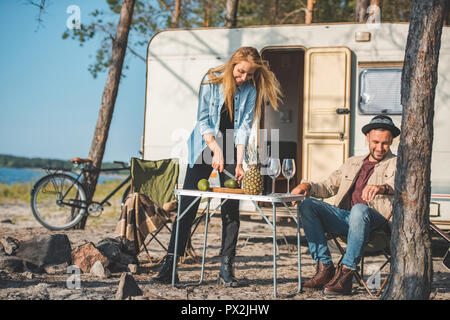 beautiful girl cutting avocados while happy man resting near in camp Stock Photo