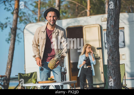 selective focus of happy man cutting pineapple while woman standing at campervan behind Stock Photo