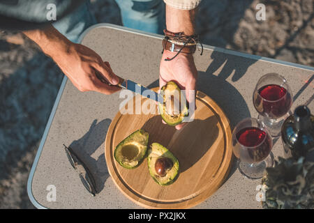 partial view of man cutting avocado on wooden board on table with wineglasses and sunglasses Stock Photo