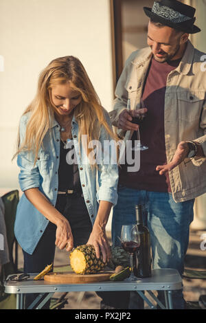 happy girl cutting pineapple while man with glass of wine talking to her Stock Photo