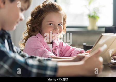 adorable little schoolgirl looking at camera while reading book with brother using tablet blurred on foreground Stock Photo