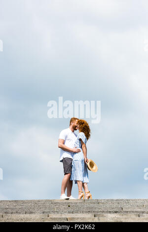 low angle view of redhead man embracing and kissing girlfriend on stairs against cloudy sky Stock Photo
