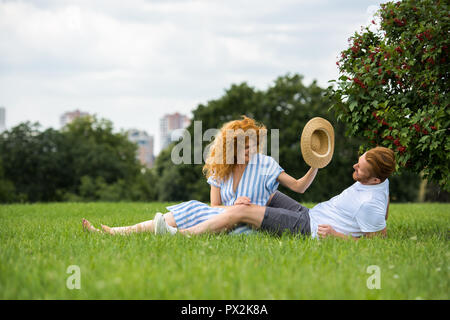 smiling redhead woman trying to putting on own straw hat on boyfriend head on grass in park Stock Photo