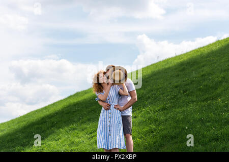smiling redhead woman covering boyfriend face by straw hat on grassy hill Stock Photo