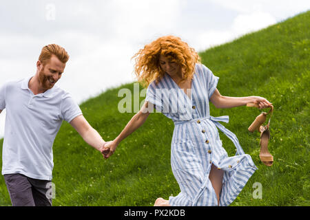 happy redhead woman holding shoes in hand and walking with boyfriend on grassy hill Stock Photo
