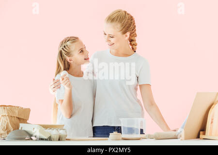 young mother and adorable little daughter in white t-shirts looking at each other during cooking isolated on pink Stock Photo