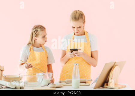 attractive young mother and daughter using smartphone while cooking together isolated on pink Stock Photo