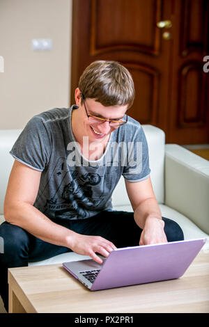 Concentrated young man with glasses working on a laptop at home office. Looking at the display and smiling Stock Photo