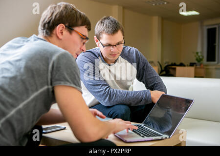 Two young people are discussing a project in the office. Sit at the table next to each other, one of them tells the other about his project and shows on the display, scrolls through the text Stock Photo