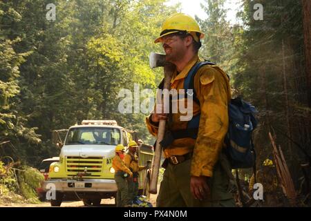 Washington Air National Guard Staff Sergeant Eric Delaune, 141st Maintenance Squadron, seen in three-quarter profile view, wearing yellow safety gear and a blue backpack, and resting an axe on one shoulder, takes a break from a clearing burnable materials while fighting the Sheep Creek Fire, with several other team members and an emergency vehicle visible in the background, located in the Sheep Creek area near Northport, Washington, USA, image courtesy Technical sergeant Timothy Chacon and the Joint Forces Headquarters, Washington National Guard, August 6, 2018. () Stock Photo