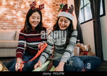 ladies sitting on the floor doing artwork together at home. young girl holding red ribbon to wrap the xmas presents. teddy bear sitting beside the wal Stock Photo