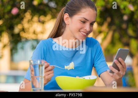 Beatutiful woman is using an app in her smartphone device, while eating a salad in an outdoor restaurant Stock Photo
