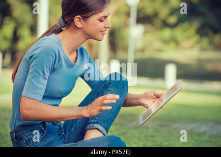 beautiful young woman is reading from a tablet pc while seating outdoors on grass in a park Stock Photo