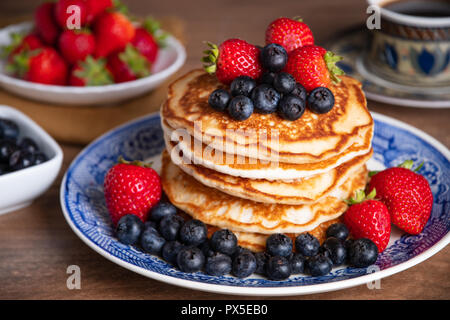 Beautiful and delicious stack of pancakes with fresh blueberries, strawberries and maple syrup on a blue and white plate with bowls of extra fruit Stock Photo
