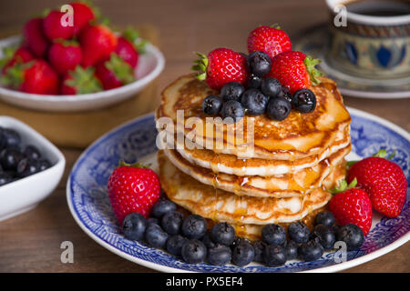 Beautiful and delicious stack of pancakes with fresh blueberries, strawberries and maple syrup on a blue and white plate with bowls of extra fruit Stock Photo