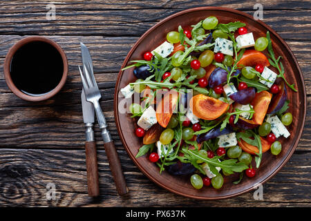 overhead view of healthy autumn salad with chocolate persimmon, blue mold cheese, green grapes, plum slices and arugula on a clay plate on an old wood Stock Photo