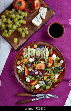 healthy autumn salad with chocolate persimmon, blue mold cheese, green grapes, plum and arugula on a clay plate with sweet vinegar dressing, vertical  Stock Photo