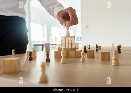 Business hierarchy concept with businessman placing chess figure of king on top of wooden stacked wooden blocks and other figures spread on office des Stock Photo
