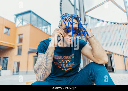 Young attractive man with blue dreadlocks talking by mobile phone and touching his hair. Stock Photo