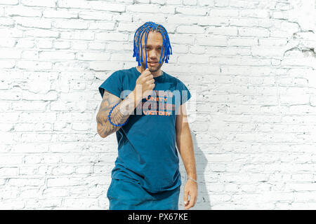 Young attractive man with blue dreadlocks touching his hair next to white bricked background. Stock Photo