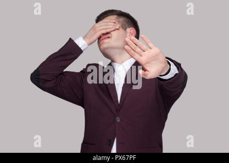 I dont want to see this. Portrait of confused young man in violet suit and white shirt, standing, closed his eyes with hands and showing stop gesture. Stock Photo