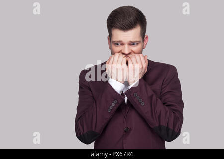 Portrait of nervous handsome young man in violet suit and white shirt, standing, looking away and bitting nails with worry face. indoor studio shot, i Stock Photo