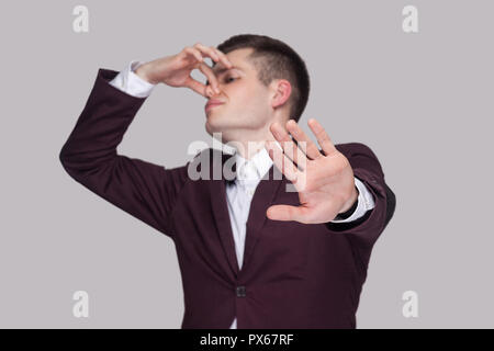 Portrait of handsome confused young man in violet suit and white shirt, standing, blocking his nose and showing stop gesture. focus on hand. indoor st Stock Photo