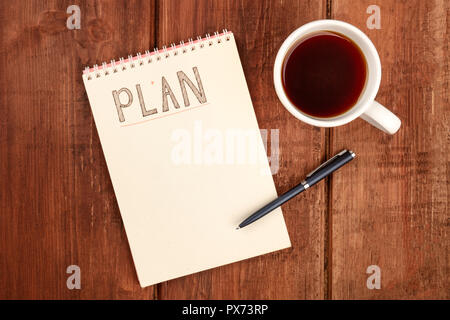A photo of a spiral notebook with the word Plan, with a cup of coffee and a pen, shot from the top on a dark rustic wooden desk with a place for text Stock Photo