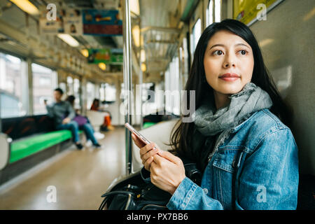 elegant lady sitting on the subway, using her cellphone and looking at the window. Asian woman traveler contemplating outdoor view from window of trai Stock Photo