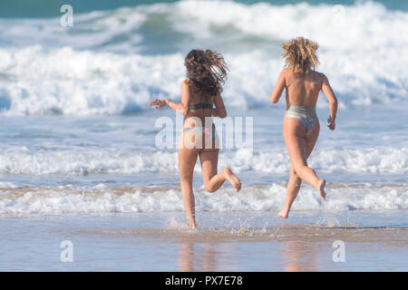 Two young attractive female holidaymakers on a staycation holiday running into the sea at Fistral in Newquay in Cornwall. Stock Photo