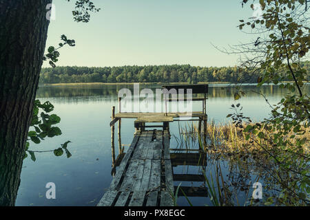 Autumn scene - lake and forest landscape. Old rotten wooden pier with missing planks with bench on a lake in the forest at autumn. Stock Photo