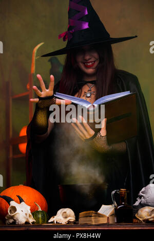 Photo of witch in black hat and with black book brewing potion Stock Photo