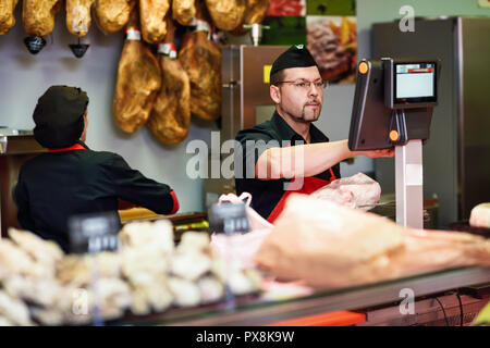 Butcher in a butcher's shop weighing the meat and charging with ham at the background Stock Photo