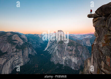 A fearless hiker is standing on an overhanging rock enjoying the view towards famous Half Dome at Glacier Point overlook in beautiful evening twilight