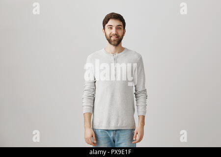 Studio shot of attractive cheerful husband in casual pullover, smiling broadly and looking at camera while standing over gray background. Client waits for barber to take care of his hair Stock Photo