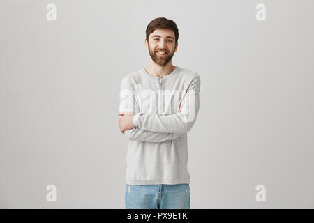 Positive shy good-looking guy with beard standing with crossed hands in pullover over gray background, smiling and feeling slightly uncomfortable of being photographed. Man does not used to attention Stock Photo