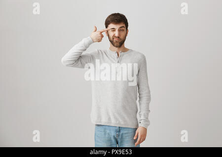 Tired or bored good-looking guy holding gun gesture near temples and making faces while standing over gray background. Upset man can not handle speaking with his annoying mother-in-law Stock Photo