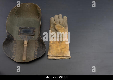 Welder's mask and gloves on a black workshop table. Protective clothing for workers in metalware. Dark background. Stock Photo