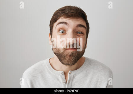 Indoor shot of attractive funny bearded guy squinting and making faces at camera, being in good mood while standing over gray background. Man mimics as if he looking through peephole Stock Photo