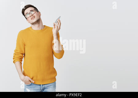 Guy bored and tired of talking with boring friend pulling face from cellphone rolling eyes and looking up suffering from annoyance and boredom cannot stand long conversations, posing over grey wall Stock Photo