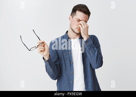 Guy rubbing eyes as feeling tired after wearing glasses while working over freelance project on computer standing with eyewear in hand and closed eyes being drained and exhausted, being hardworking Stock Photo