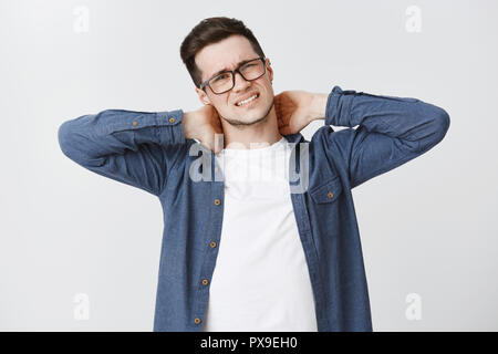 Waist-up shot of displeased young man feeling discomfort in neck touching back with hands frowning and clenching teeth from pain having problems with spine as sitting too long in front of computer Stock Photo