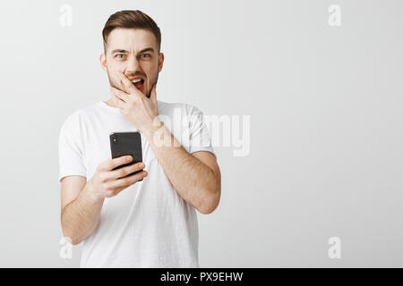 Shocked stunned handsome guy opening up terrible photo of ex-girlfriend shouting from dislike and surprise covering opened mouth and frowning staring intense holding smartphone in hand Stock Photo