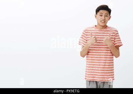 Fine I guess. Portrait unsure awkward young attractive asian man in striped t-shirt making tight ucertain smile and showing thumbs up gesture as if agree or like idea, posing gray background Stock Photo - Alamy