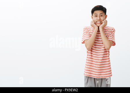 Cuteness overload. Portrait of silly attractive nice asian guy with dark short hair smiling broadly leaning face on palms in glamorous feminine pose posing in striped t-shirt against white background