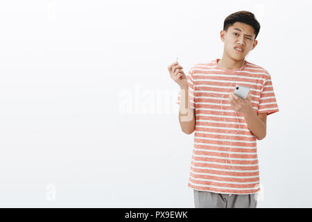 Earphones broke down, guy disturbed with awful sound, taking off earbud frowning and turning away from headphone. Asian cute guy holding smartphone looking bothered and displeased at camera Stock Photo