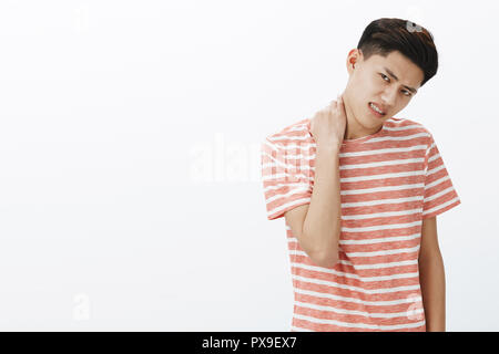 Portrait of bothered uneasy young asian man in striped t-shirt unwilling to do something rubbing neck tilting head and frowning expressing displeasure and awkward feeling over white background Stock Photo