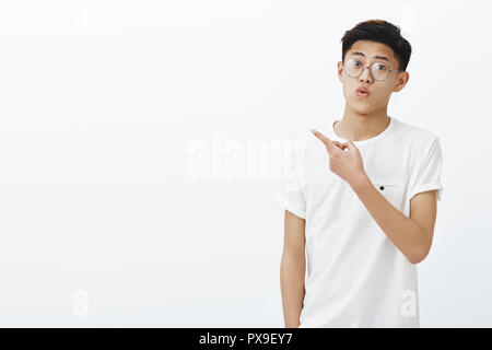 Curious and impressed young stylish handsome asian boy in round glasses folding lips, making wow sound from excitement and amazement pointing at upper left corner, expressing surprise and interest Stock Photo
