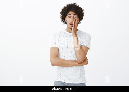 Guy sympathizing friend, being shocked while hearing terrible story. Impressed and amazed good-looking hispanic male coworker in t-shirt, dropping jaw, touching cheek and staring astonished at camera Stock Photo